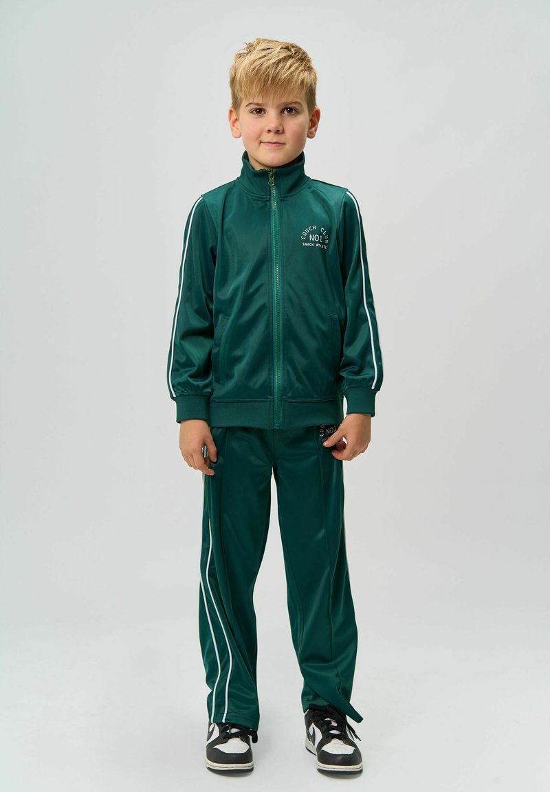 Tuta verde in poliestere con strisce bianche, giacca con zip e pantaloni con vita elastica. Sneakers nere e bianche completano l'outfit.