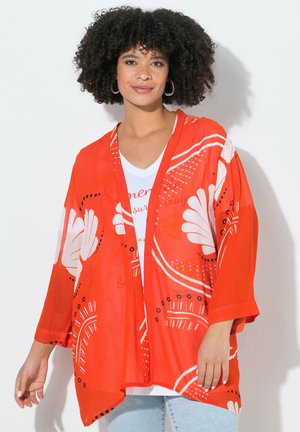 Mujer con cabello rizado que lleva pendientes grandes de aro, una chaqueta estilo kimono de color naranja brillante con estampado sobre una camisa blanca y vaqueros azul claro.
