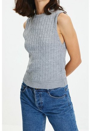 Vrouw draagt een mouwloze grijze kabelgebreide trui, gestopt in een blauwe high waisted jeans, handen achter de rug, witte achtergrond.