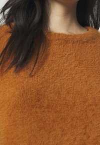 Donna con lunghi capelli scuri che indossa un maglione morbido di lana arancione con scollatura rotonda, inquadrata dal collo fino a metà torace.