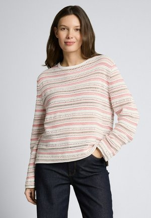 Vrouw die een lichtroze en wit gestreepte gebreide trui en donkerblauwe jeans draagt, staand met één hand in de zak tegen een effen achtergrond.