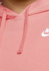 Rosa hoodie med en strukturerad yta, med en luva med dragsko och en vit Nike-logotyp. Närbild som framhäver tygdetaljer.