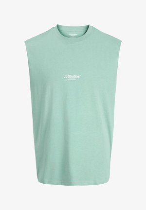 Piedurkņu zaļa krekla bez piedurknēm ar apaļu kakla izgriezumu, uz kura priekšpuses centrā ir uzdrukāts balts "JJ Studios" logotips un neliels teksts.