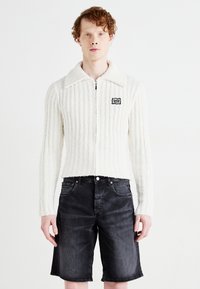 Suéter blanco con cremallera y canalé, con un gran cuello y parche de logo negro, combinado con pantalones cortos de mezclilla oscuros. Textura suave y diseño ajustado.