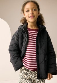 Veste matelassée noire avec une capuche, sur un pull rayé bordeaux. Associée à un pantalon crème à imprimé léopard, avec des poches et une fermeture éclair.