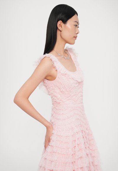 Halvány rózsaszín fodros ruha texturált rétegekkel, négyzet alakú nyakkivágással és áttetsző anyagból. A modell virágos díszítésű nyakláncot visel.