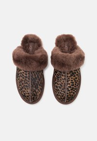 Pantofole con stampa leopardata, foderate e bordate in pelliccia sintetica marrone. Forma arrotondata con dettagli cuciti. Texture morbida e aspetto accogliente.
