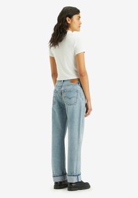 Lichtblauwe high-waisted jeans met een rechte pasvorm, zichtbare stiksels en een bruine leren patch op de achterkant, gecombineerd met een witte top en zwarte schoenen.
