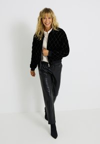 Veste bomber en velours noir avec motif de matelassage en losanges, poignets et taille en tricot, associée à un pantalon en faux cuir noir et des bottines pointues.