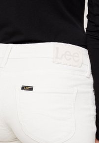 Person som bär vita kordbyxor med Lee-märket på bakfickan och midjan, i kombination med en svart långärmad topp.