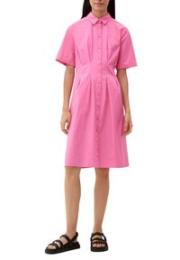 Różowa sukienka koszulowa z krótkim rękawem, z zapięciem na guziki, plisowanym fasonem i prostym dołem. Noszona z czarnymi sandałami na cienkich paskach.