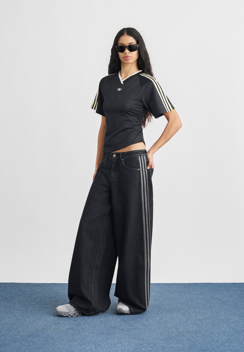 Frau trägt schwarzes Adidas-Shirt mit goldenen Streifen, weite schwarze Hose mit silbernen Seitenstreifen, weiße Turnschuhe und schwarze Sonnenbrille, steht auf blauem Boden.