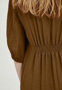 Robe en tissu brun texturé avec des manches bouffantes et des détails de taille froncée. Le matériau semble léger et légèrement froissé.