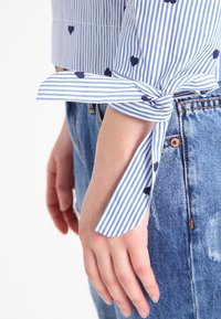 Blusa a righe blu e bianche con motivi a cuori blu navy. Presenta maniche annodate, abbinata a un tessuto in denim di jeans a vita alta.