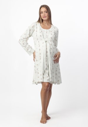 Mujer de pie descalza con un vestido blanco hasta la rodilla, de manga larga y con un pequeño estampado floral, posando con una mano en la cadera.