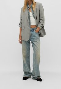 Femme portant un blazer gris clair sur une veste en jean et un crop top blanc, avec un jean bleu délavé ample et des chaussures noires, debout avec la main dans la poche.