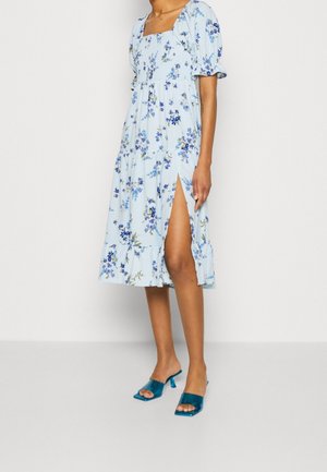 Femme portant une robe midi bleu clair à fleurs avec manches ballons et une haute fente latérale, associée à des sandales à talons ouvertes couleur sarcelle.