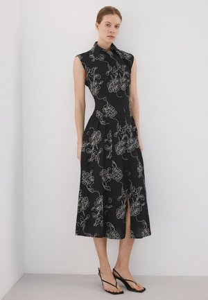Femme portant une robe midi noire sans manches avec broderie florale blanche, boutonnée devant, col et fente avant, associée à des sandales noires à talons.