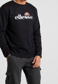 Sweat-shirt noir avec poignets côtelés, présentant un grand logo blanc "ellesse" et un graphique coloré de demi-soleil sur la poitrine. Design simple et décontracté.