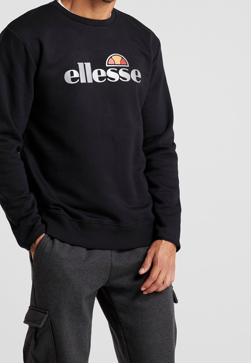 Sweat-shirt noir avec poignets côtelés, présentant un grand logo blanc "ellesse" et un graphique coloré de demi-soleil sur la poitrine. Design simple et décontracté.