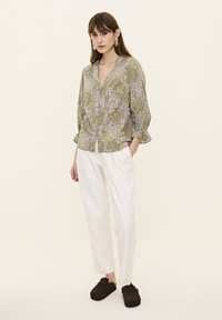 Grön blus med blommönster, lång ärm och V-ringning, kombinerad med vita, löst sittande byxor och mörkbruna slip-on skor.