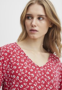 Rote Bluse mit V-Ausschnitt, verziert mit kleinen weißen und schwarzen Blumen auf einem strukturierten Hintergrund. Kurze Ärmel und fließendes Design.