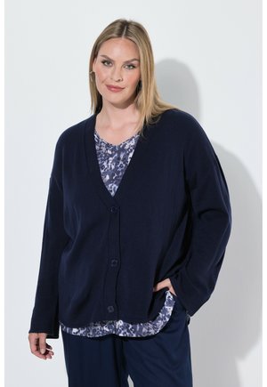 Lyshåret kvinde iført en mørkeblå knappet cardigan over en mønstret bluse, med den ene hånd i lommen, stående mod en lys baggrund.