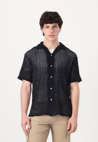 Camisa negra de punto abierto, de manga corta y con cuello, que presenta un cierre frontal con botones y un patrón texturizado en zigzag en toda la prenda.