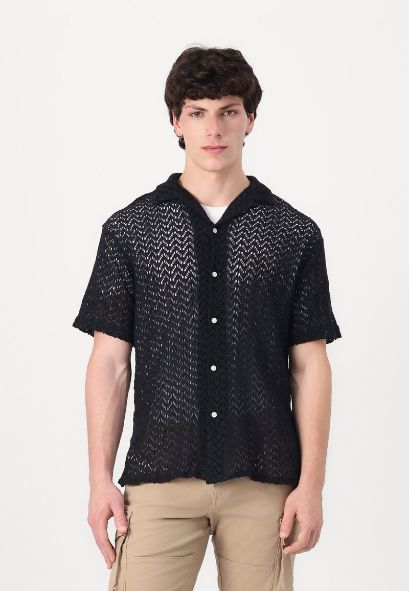 Camisa negra de punto abierto, de manga corta y con cuello, que presenta un cierre frontal con botones y un patrón texturizado en zigzag en toda la prenda.