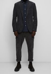 Blazer in velluto a coste grigio scuro con un design strutturato, abbinato a una camicia button-up blu navy. Pantaloni cargo grigio scuro e stivaletti neri.