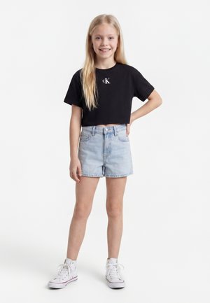 Fille souriante aux longs cheveux blonds portant un t-shirt noir court Calvin Klein, un short en jean clair et des baskets blanches montantes, debout la main sur la hanche.