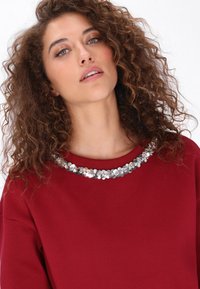 Sudadera color granate con cuello redondo adornado con lentejuelas plateadas. Textura suave con un ajuste relajado.