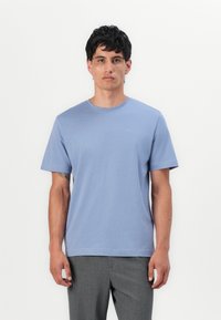 Hellblaues Baumwoll-T-Shirt mit Rundhalsausschnitt, kurzen Ärmeln und einem dezenten Logo auf der Brust. Kombiniert mit grauen Hosen.