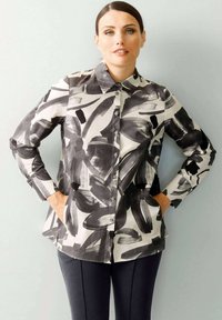 Weißes Hemd mit einem abstrakten Pinselstrichmuster in Schwarz. Lange Ärmel, klassischer Kragen und Seitentaschen; glatte Textur.