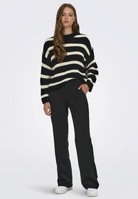 Czarny i kremowy dzianinowy sweter w paski o luźnym kroju, zestawiony z czarnymi spodniami i białymi trampkami, na neutralnym tle.