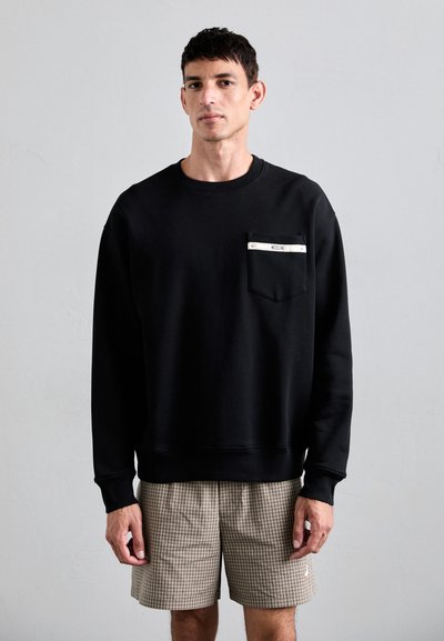 Czarny sweatshirt z okrągłym dekoltem, wyposażony w kieszeń naszywaną z metalicznym akcentem. Noszony z beżowymi, w kratkę szortami. Miękka faktura.