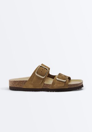 Bruine suède sandaal met open hiel, twee verstelbare zilveren gespen en een kurken zool op een witte achtergrond.