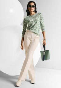 Frau in grün gemustertem Pullover, mit cremfarbenen Hosen und weißen Stiefeln, trägt grüne Sonnenbrille und hält eine grüne Handtasche, steht vor einem weißen kreisförmigen Hintergrund.