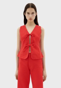 Red linen sleeveless top with a V-neck and front bow ties, featuring a fitted shape and subtle peplum hem. Paired with matching trousers.