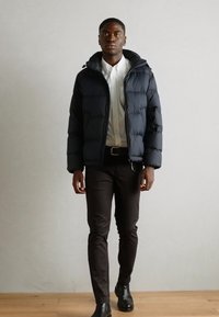 Doudoune marine avec zip et capuche, associée à une chemise blanche et un pantalon sombre ; le modèle se tient sur un sol en bois.