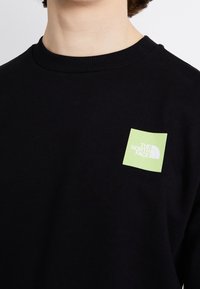 Persona vistiendo un suéter negro de cuello redondo con un pequeño logo cuadrado de color verde claro que dice "The North Face" en la parte superior del pecho.