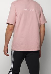 Vaaleanpunainen puuvillapaita, jossa on lyhyet hihat ja pyöreä kaula-aukko. Paidassa on pieni valkoinen logo selässä. Yhdistetty mustiin housuihin, joissa on valkoiset raidat.