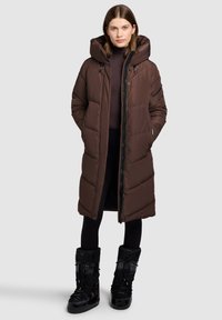 Ruskea toppatakki hupulla, jossa vetoketjullinen etuosa ja sivutaskut. Päällä tumma pooloneule ja mustat saappaat.