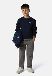 Felpa navy con logo rotondo, pantaloni grigi e sneakers beige. Il modello tiene in mano una giacca navy trapuntata con fodera verde. Vestibilità casual.