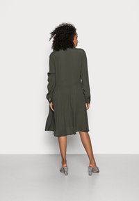 Vestido verde oliva com uma saia fluida, mangas longas e cintura ajustada. Combinado com sandálias de salto bloco xadrez. Textura de tecido suave.