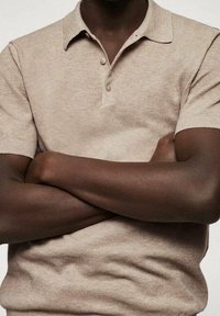 Polo beige avec un col, à manches courtes et deux boutons au cou ; texture tricot fine ; porté avec les bras croisés.