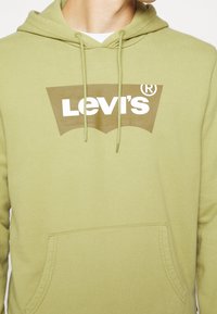 Homme portant un sweat à capuche vert clair avec un grand logo Levi's marron et des cordons, poche avant visible.