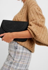 Žena v objemném béžovém svetr s copánkovým vzorem a kostkované sukni, držící velkou černou surovou koženou kabelku u boku.