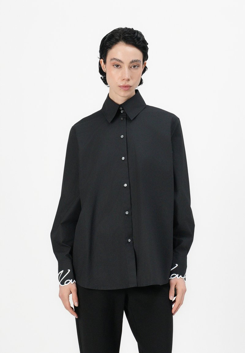 Camicia nera a maniche lunghe con colletto strutturato, dotata di chiusura con bottoni sul davanti e testo ricamato sui polsini. Tessuto in cotone morbido.