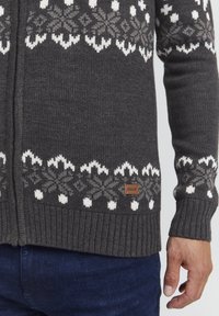 Grau gestrickte Strickjacke mit einem weißen und grauen Schneeflockenmuster, gerippten Bündchen und Saum, ausgestattet mit einem kleinen braunen Etikett mit der Aufschrift "ISOLID."